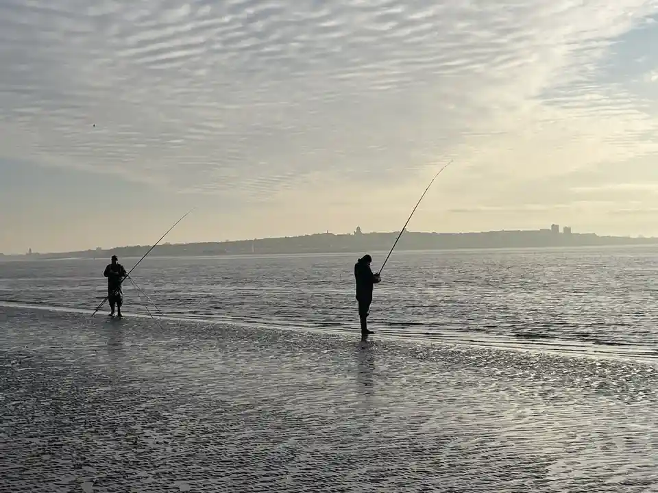 Two silhouetted people stand in shallow water on a beach, each holding a bent fishing rod, with a distant city skyline under…