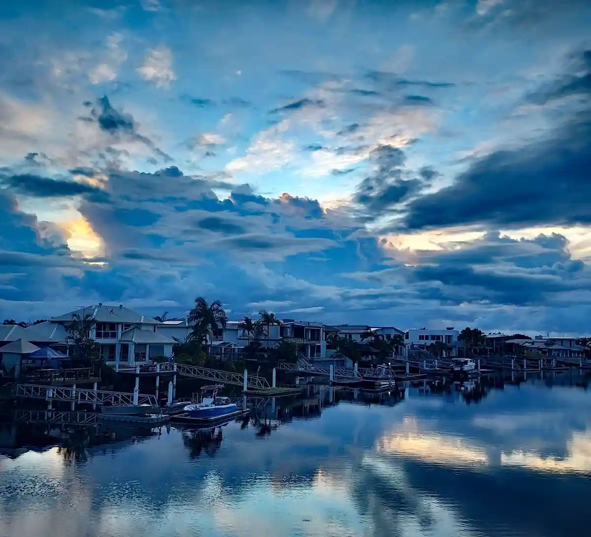 A serene waterfront scene at dusk with houses along a calm canal, boats docked, and dramatic clouds reflecting in the water…