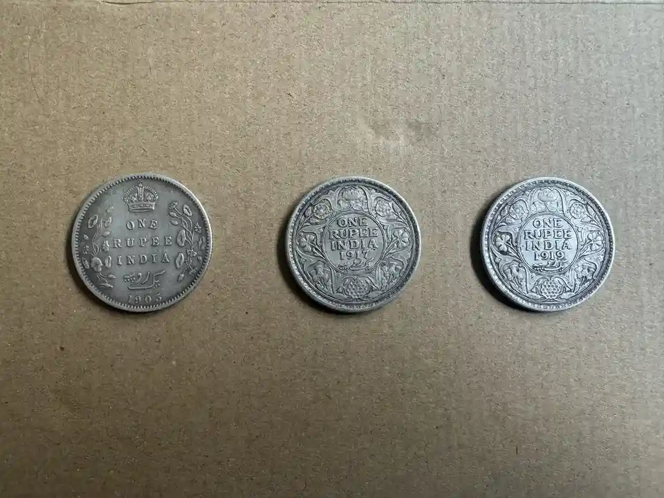 Three silver Indian one rupee coins from 1903, 1917, and 1919 are laid out side-by-side on a brown paper surface. The…