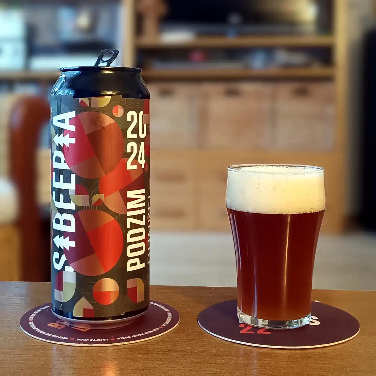 A black beer can labeled 'SBEERA 2024 PODZIM' sits next to a glass of dark amber beer with a white head, both on coasters on…
