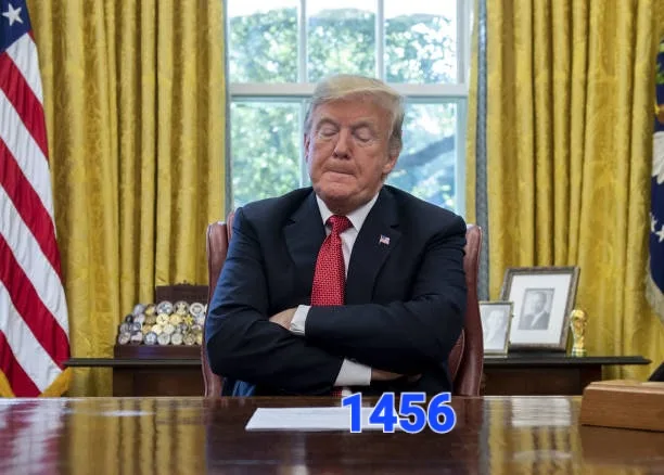 A man with blond hair, wearing a dark suit and red tie, sits at a large wooden desk with arms crossed and eyes closed in the…