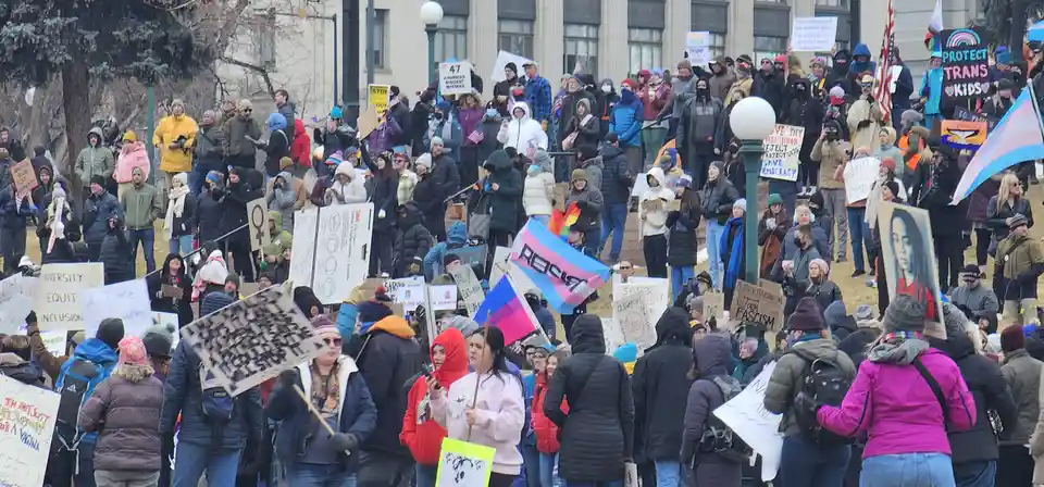 A large crowd of protesters gathered outdoors in winter, holding signs and flags including a prominent trans flag with…