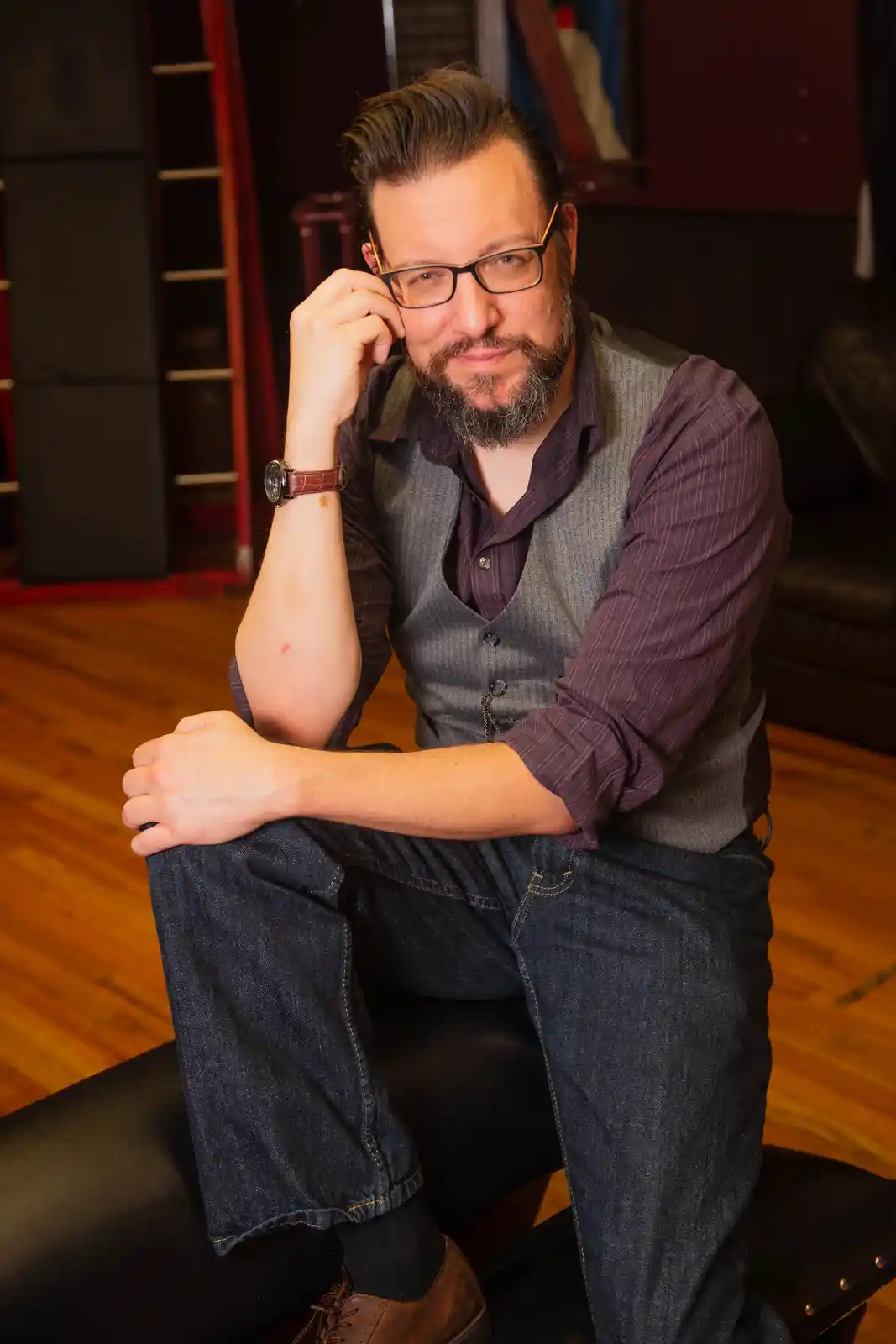 A man with glasses and a beard sits on a black leather bench, wearing a purple shirt, grey vest, and jeans. He rests his…