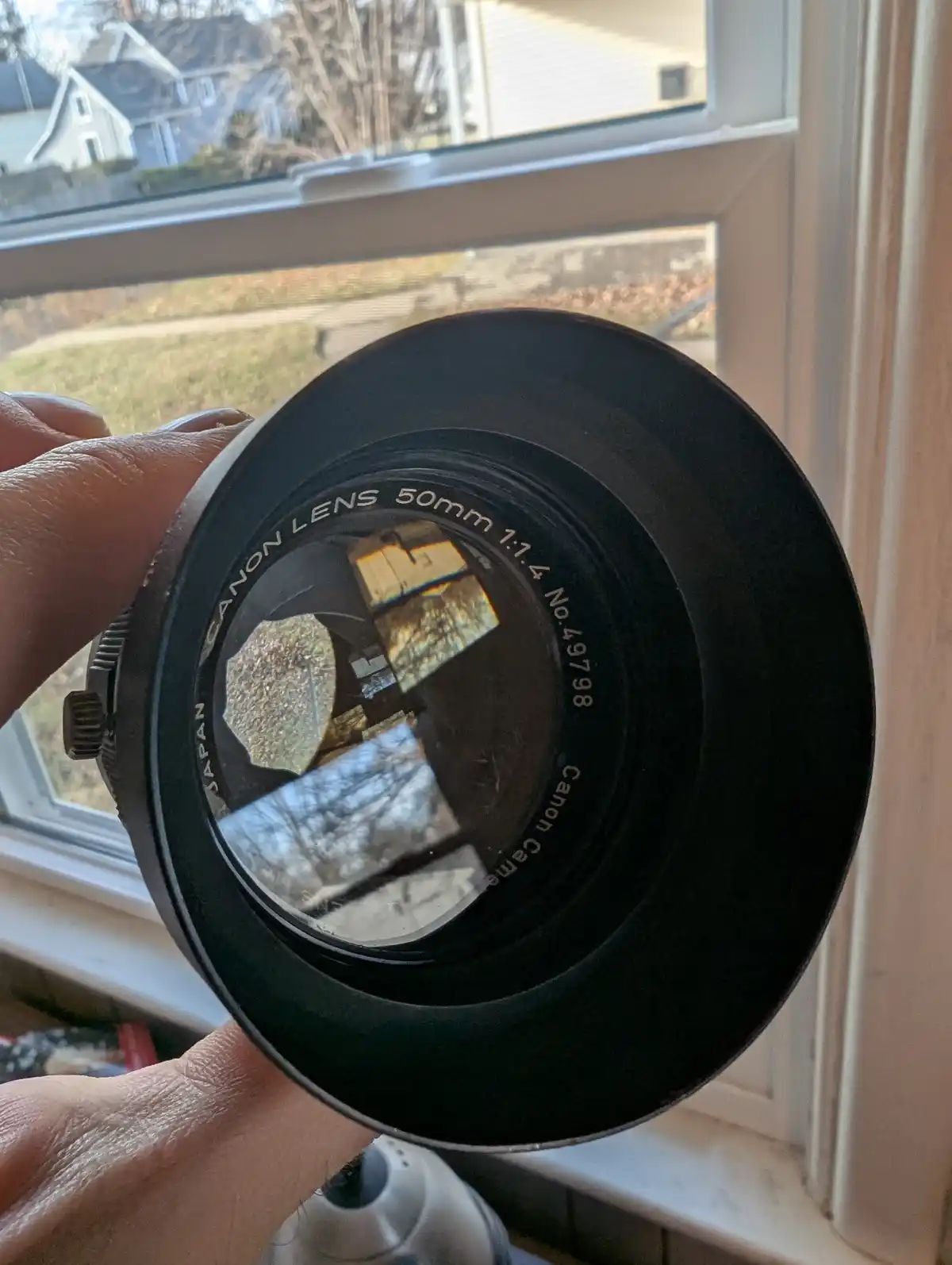 A hand holds a black Canon 50mm f/1.4 lens directly in front of a window, showing the lens's aperture blades and the…