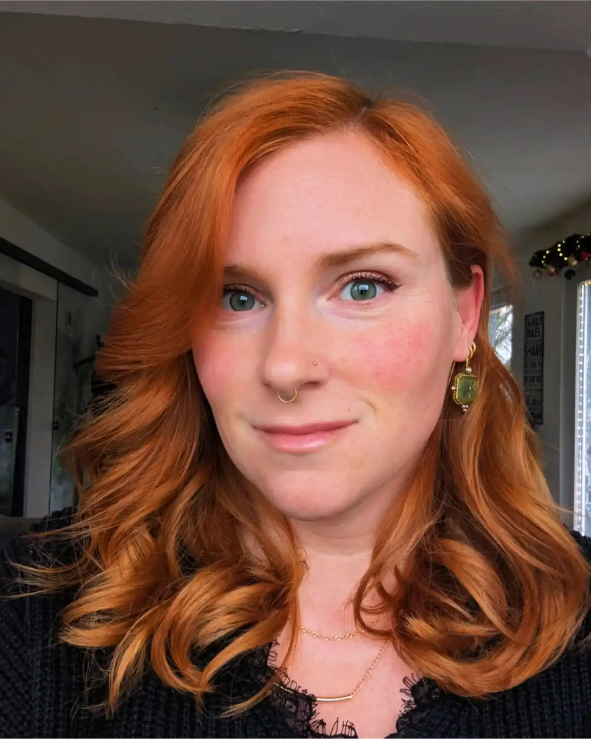 A woman with vibrant red-orange wavy hair, green eyes, and a nose ring looks directly at the camera with a subtle, slightly…