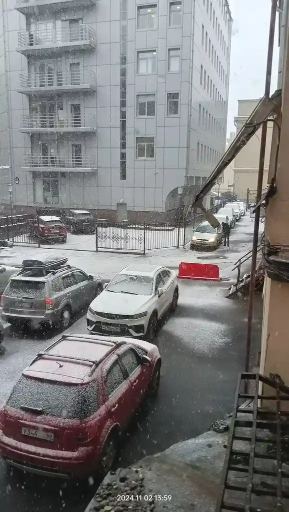A snowy urban street scene viewed from a window, showing parked cars covered in snow, a multi-story building, and a person…
