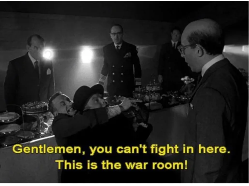 A black-and-white still from a film shows several men in suits in a dimly lit room with a long table covered in food and…