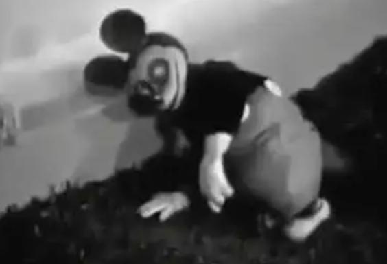 A black-and-white, grainy image shows a distorted, unsettling version of Mickey Mouse crouching on grass. His face is…