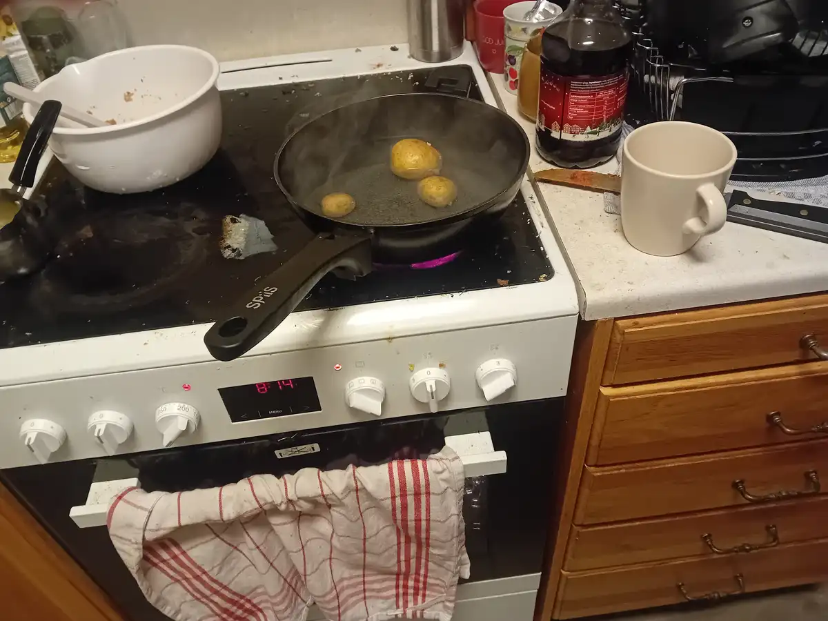 A messy kitchen stove shows a frying pan with three small potatoes sizzling in a small amount of oil, with steam rising. The…