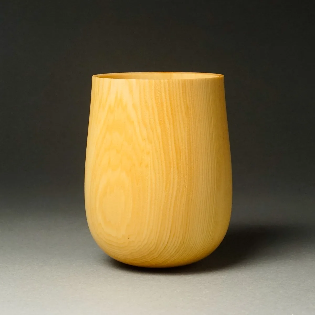 A smooth, light-colored wooden cup with vertical grain patterns sits centered on a gray surface against a dark gray…