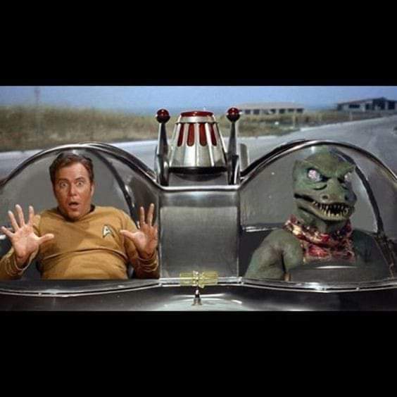 A man in a Starfleet uniform sits in the driver's seat of a futuristic car, hands raised in panic, while a green reptilian…