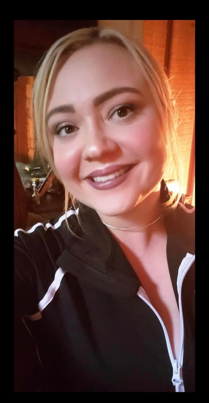 A selfie of a smiling woman with blonde hair wearing a black zip-up jacket with white stripes, taken indoors with warm…