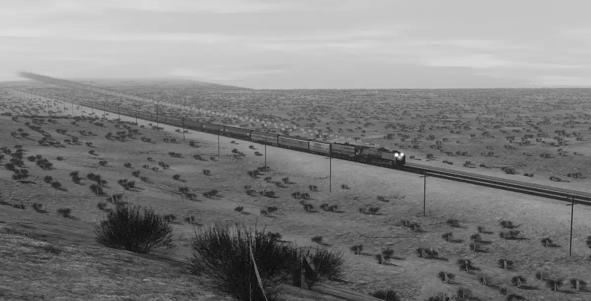 A black-and-white image shows a long freight train traveling across a vast, arid, open landscape with sparse vegetation and…