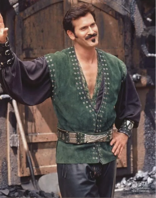 A man with a mustache wearing a green studded vest over a dark shirt and leather pants, standing in front of wooden…