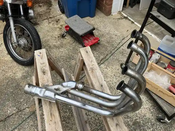 A motorcycle exhaust system with multiple curved pipes rests on wooden blocks in a garage or driveway, with the front wheel…