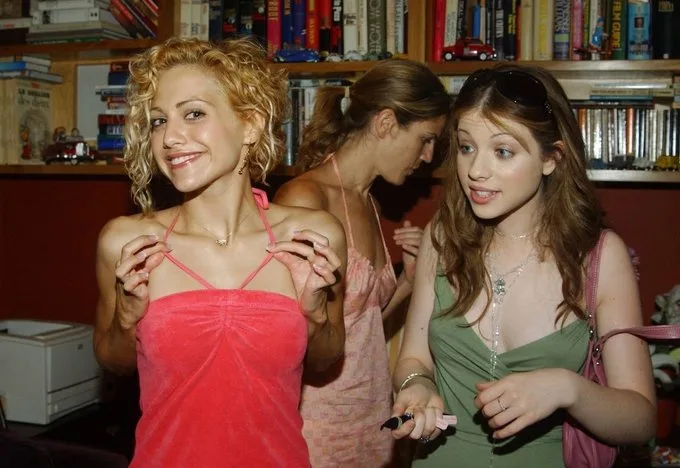 Three young women are in a room with bookshelves in the background. The woman on the left, with curly blonde hair and a pink…