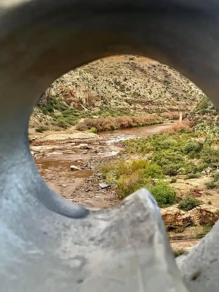 A photograph taken from inside a large, circular metal pipe or tunnel, looking out onto a winding river flowing through a…
