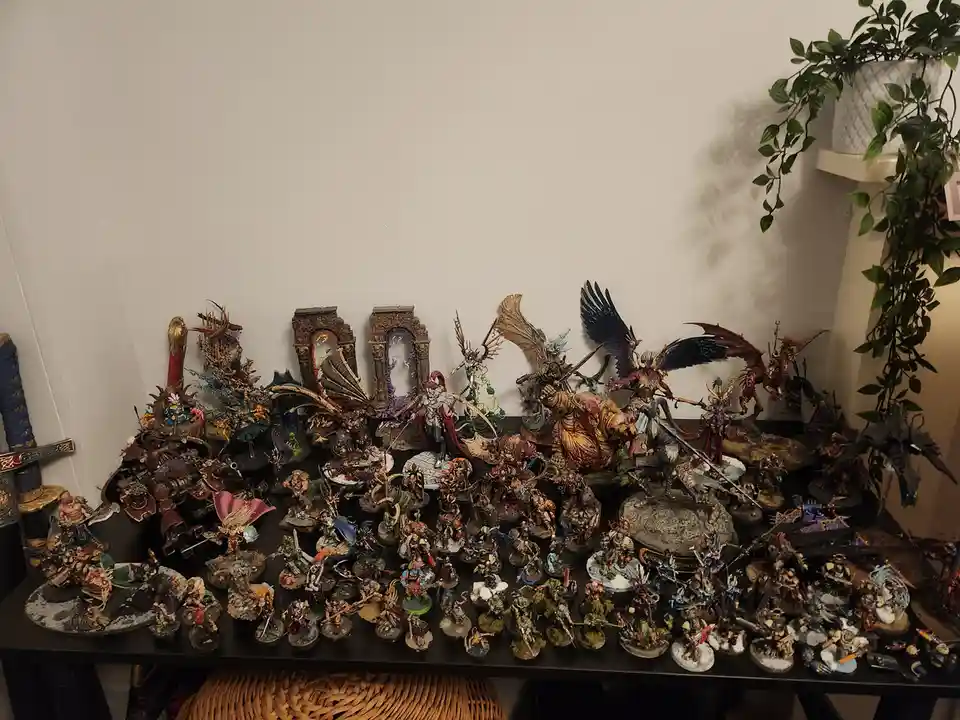 A black table is covered with hundreds of intricately painted fantasy miniatures, including large winged figures, armored…