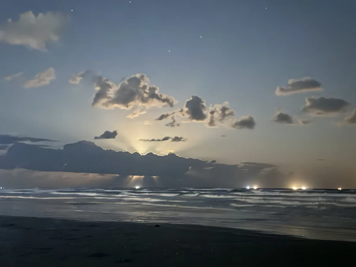A serene beach scene at dusk or dawn with scattered clouds, visible stars, and gentle waves. The horizon shows distant…