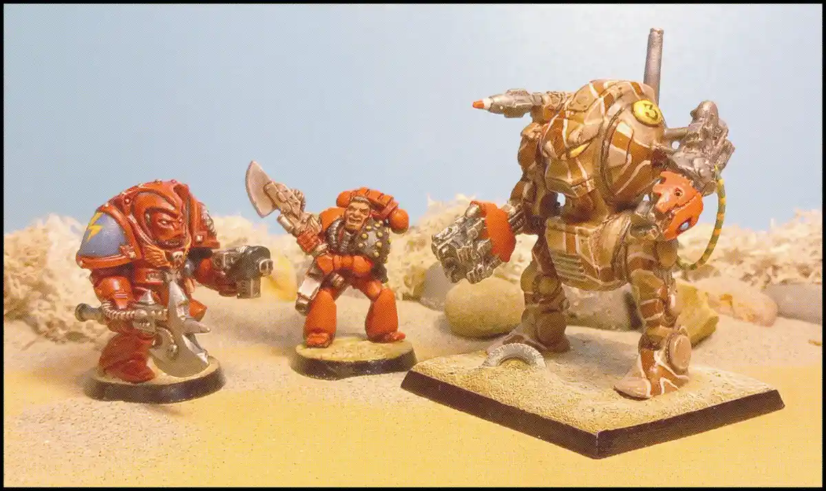 Three painted miniature figures from a tabletop wargame stand in a sandy diorama. The figure on the right, a large heavily…