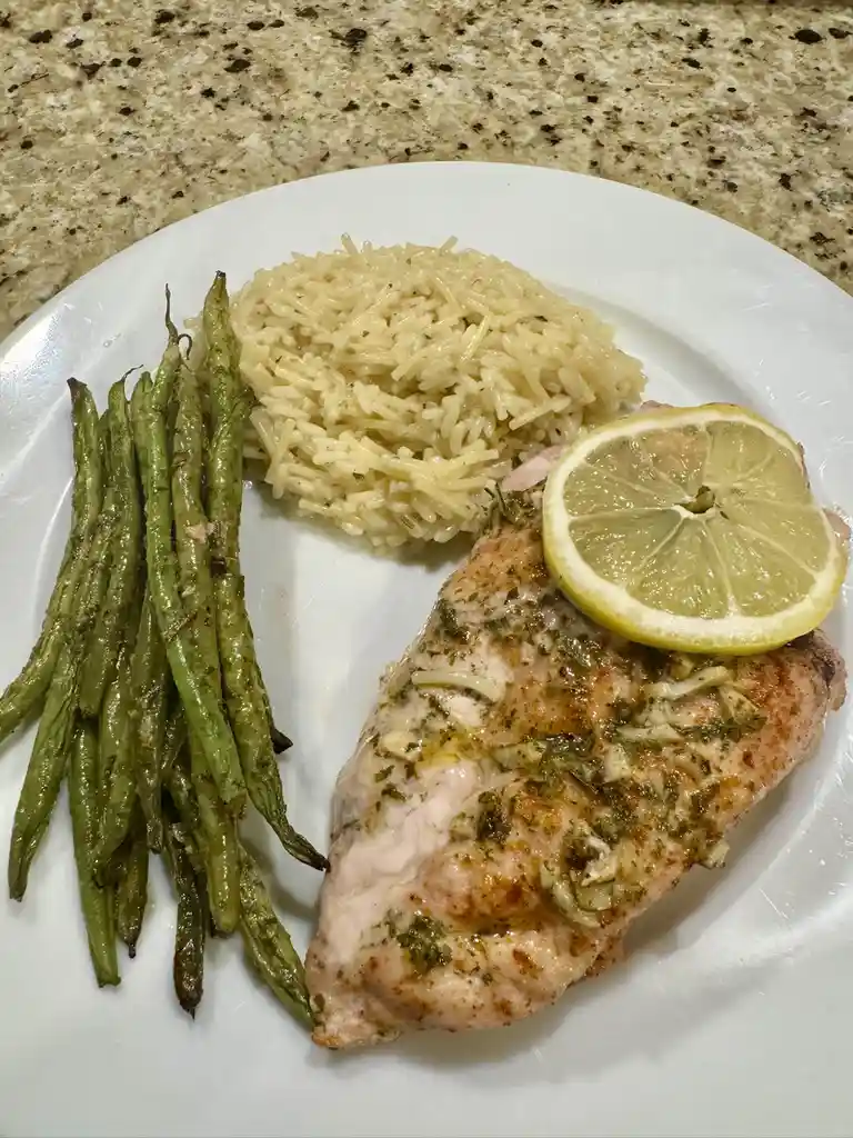 A white plate holds a cooked chicken breast topped with a lemon slice, a serving of rice, and a side of roasted green beans…