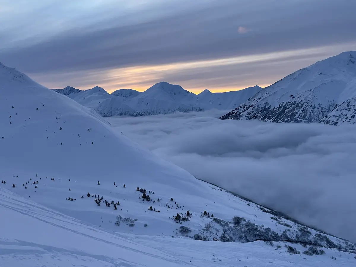 A breathtaking snowy mountain landscape at dawn or dusk, with peaks silhouetted against a soft orange and purple sky. A…