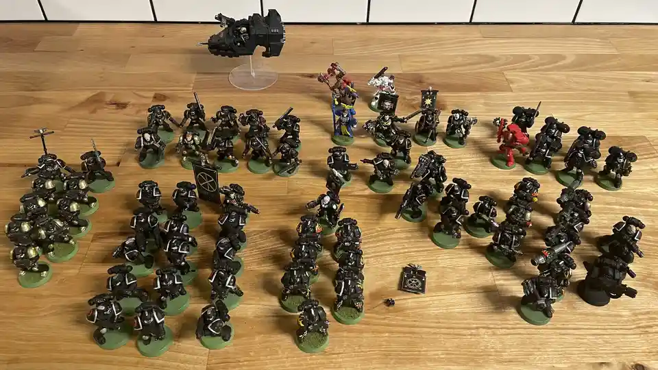 A large collection of painted miniature warhammer 40k space marine figures arranged on a wooden floor, with a single black…