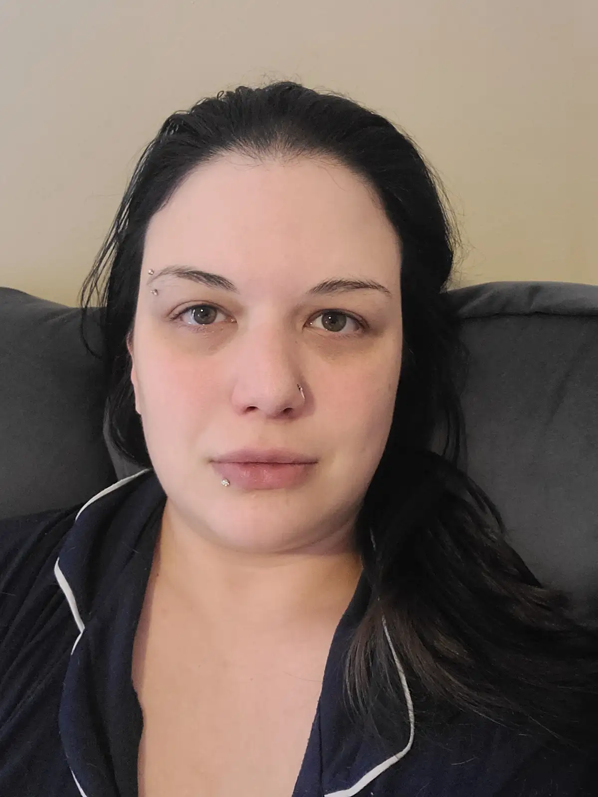 A person with dark hair and multiple facial piercings — including eyebrows, nose, and lip — stares directly at the camera…