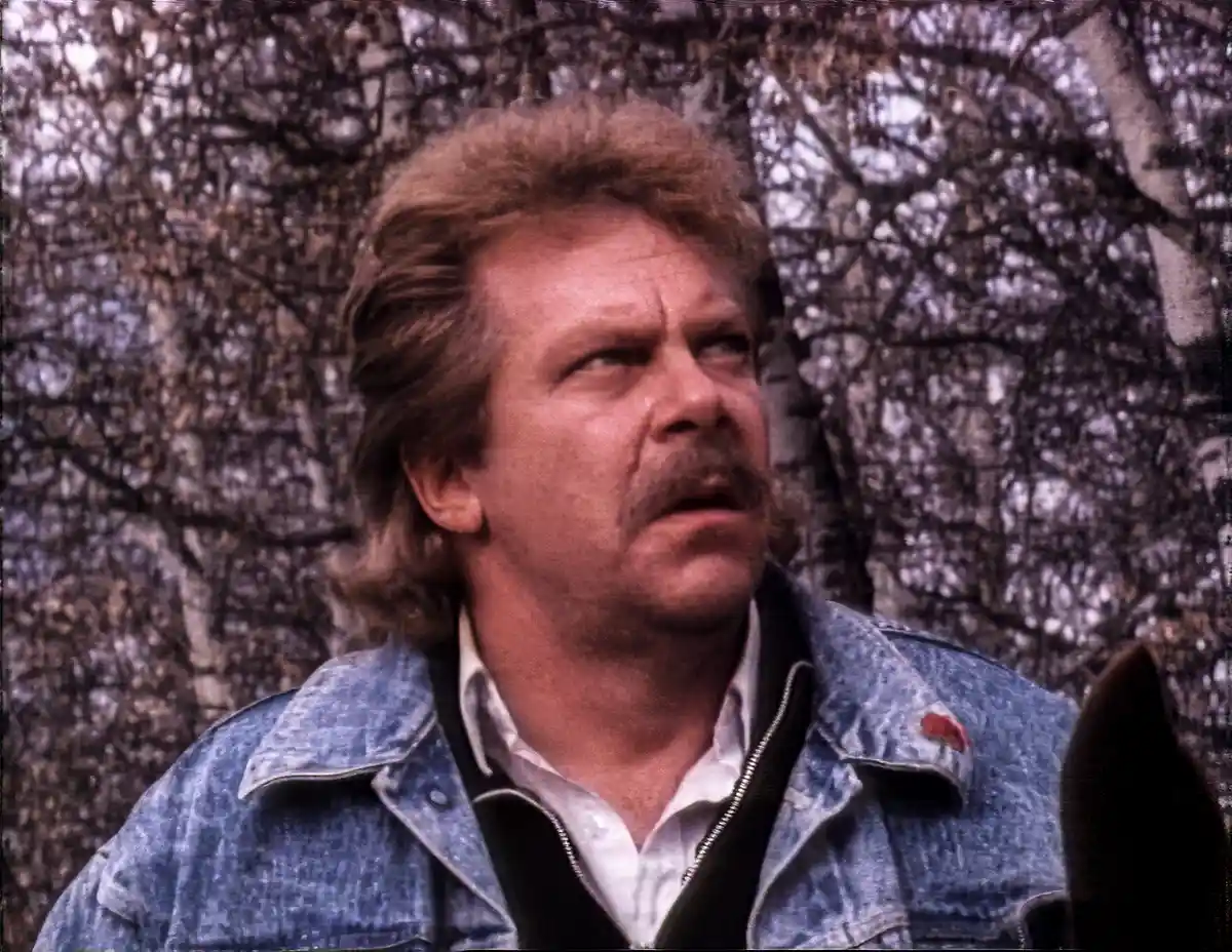 A man with a mustache and shaggy hair, wearing a denim jacket over a white shirt, looks off to the side with a skeptical…