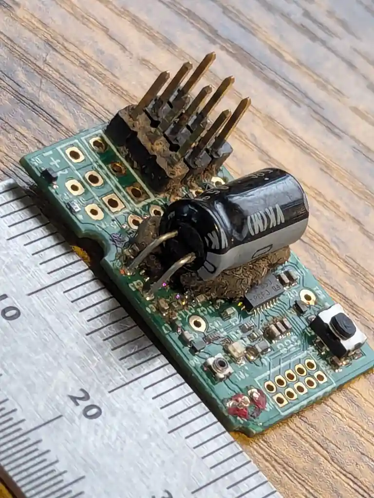 A close-up photo shows a damaged green circuit board with a burnt-out black capacitor and melted solder, resting on a wooden…