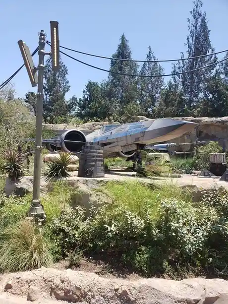 A weathered, blue-and-gray X-wing starfighter from Star Wars is displayed outdoors on a rocky, grassy terrain with trees and…