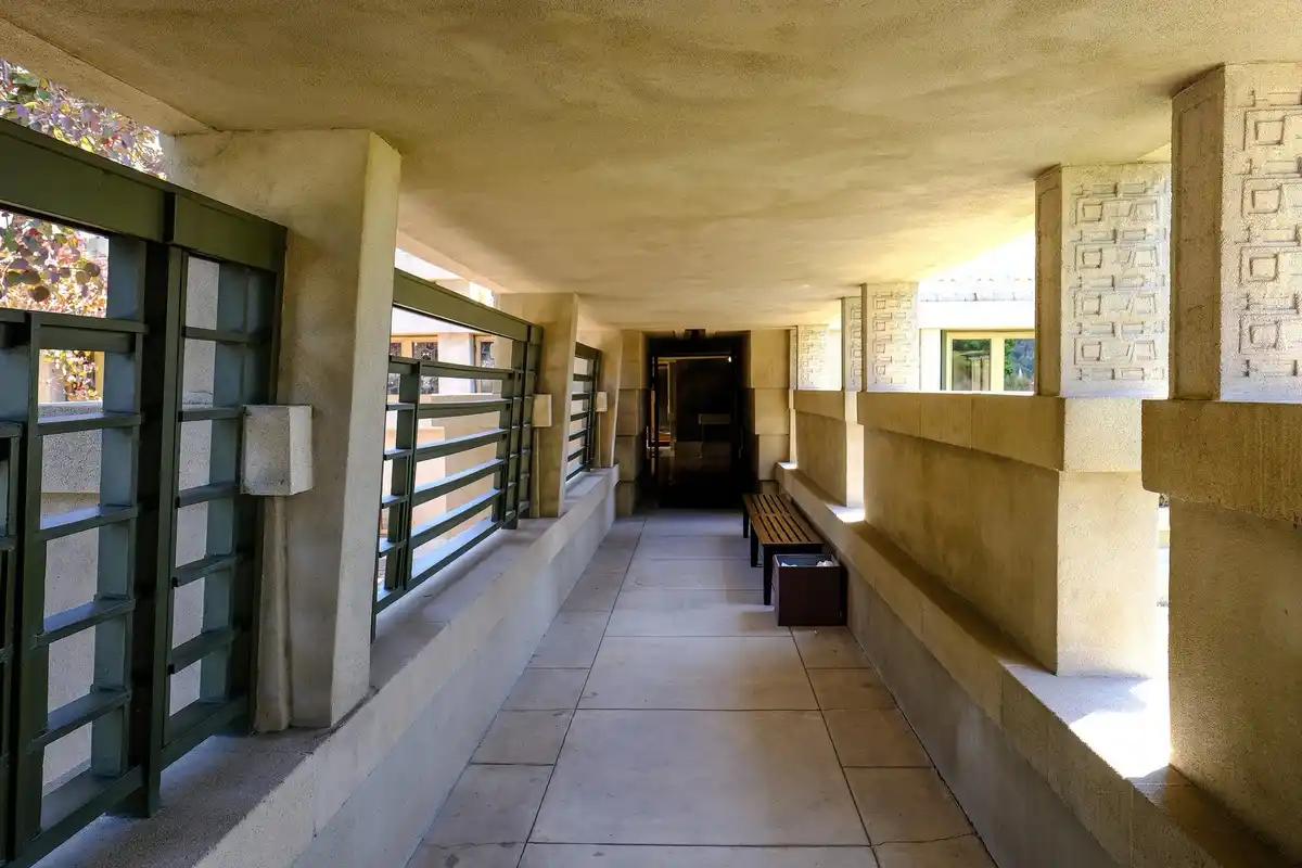 A long, sunlit concrete hallway with geometrically patterned walls and green metal railings, featuring a wooden bench and a…