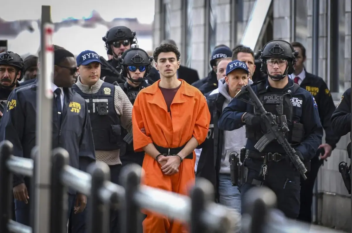 a man in an orange jumpsuit stands handcuffed, surrounded by heavily armed NYPD and FBI officers in tactical gear. The…