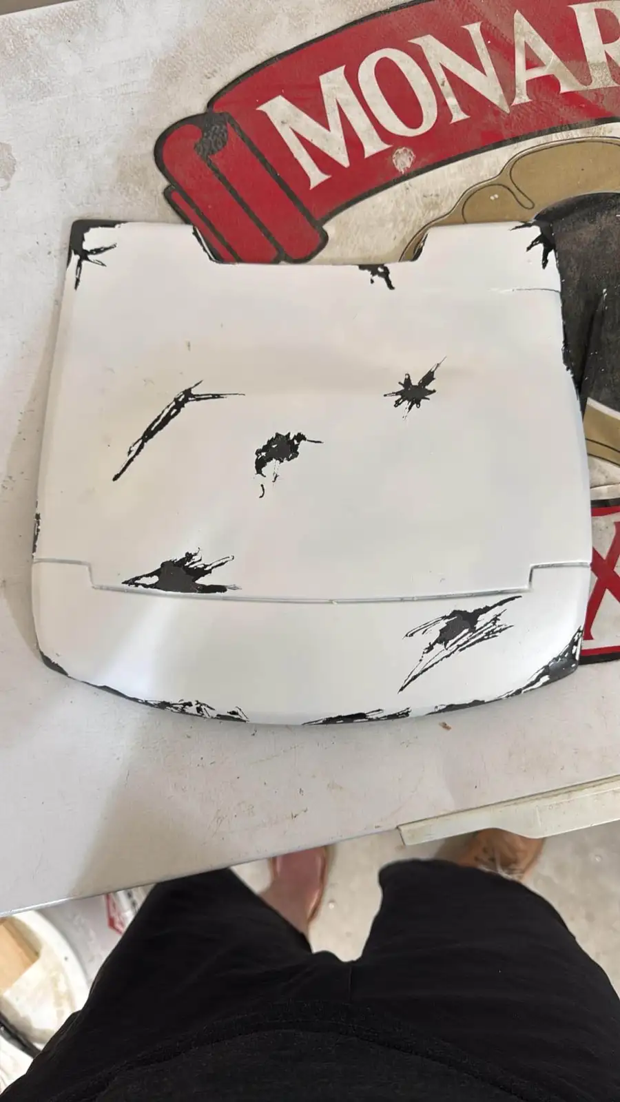 A white car hood with multiple black paint splatters and drips across its surface, resembling damage or a chaotic painting…