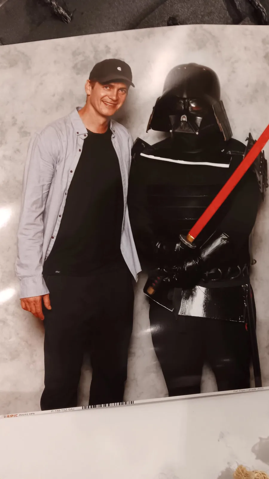 A printed photo shows actor Mark Hamill smiling beside a Darth Vader cutout. The cutout has been crudely modified with a red…