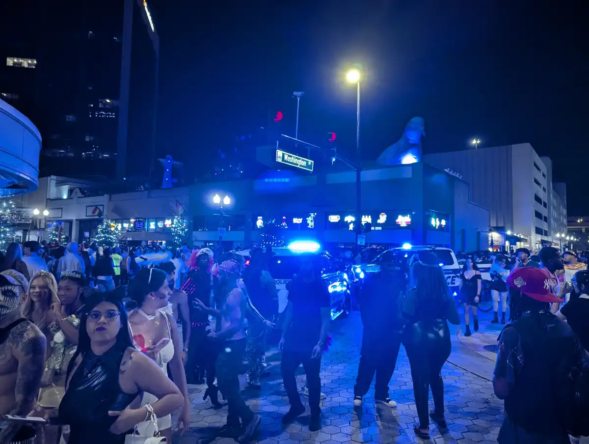 A nighttime street scene in a crowded urban area, likely a gay pride or festival event, with many people in costumes and…