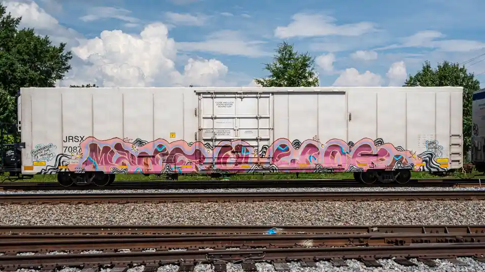 A white freight train car sits on tracks under a blue sky with clouds, covered in vibrant pink and purple graffiti. The text…