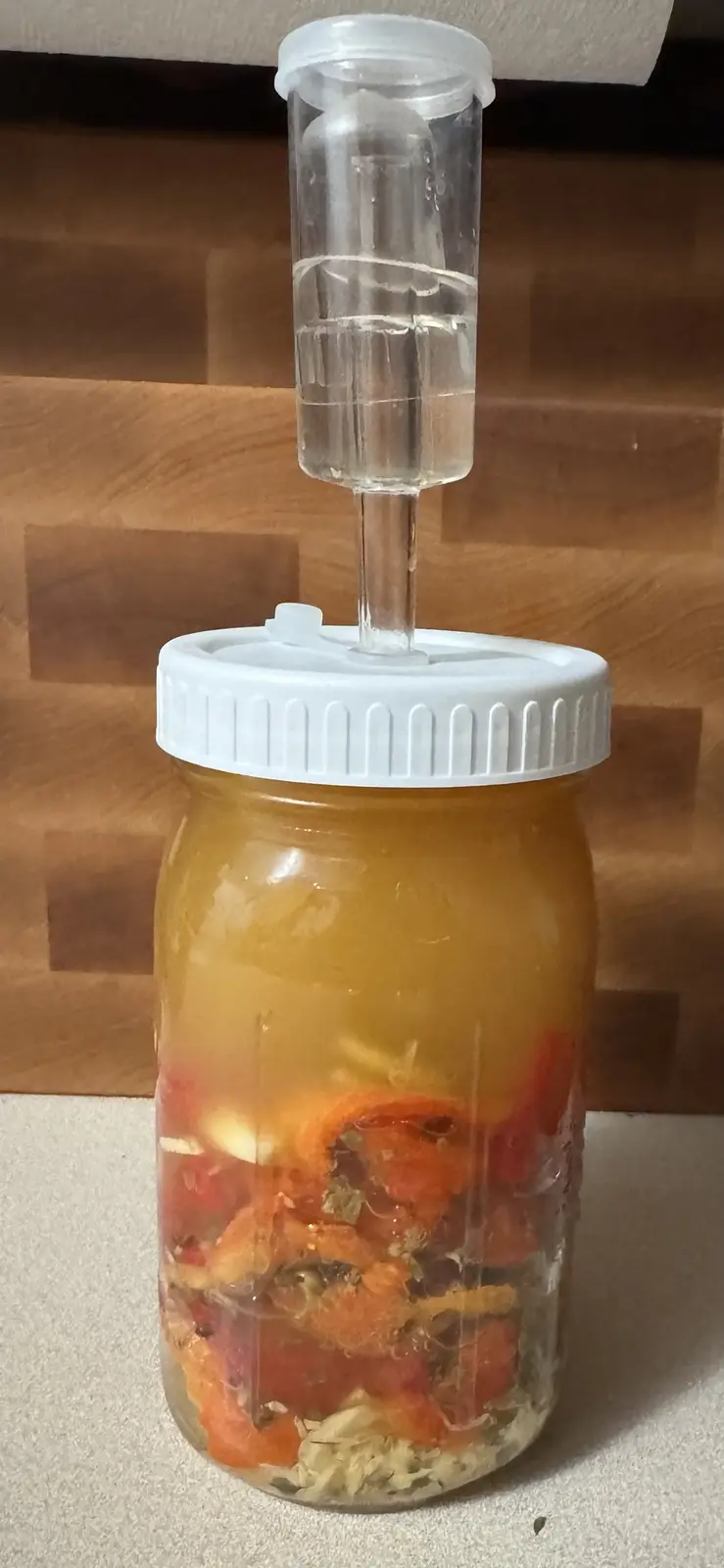 A glass mason jar filled with orange liquid and chopped red peppers sits on a countertop, topped with a white lid and a…