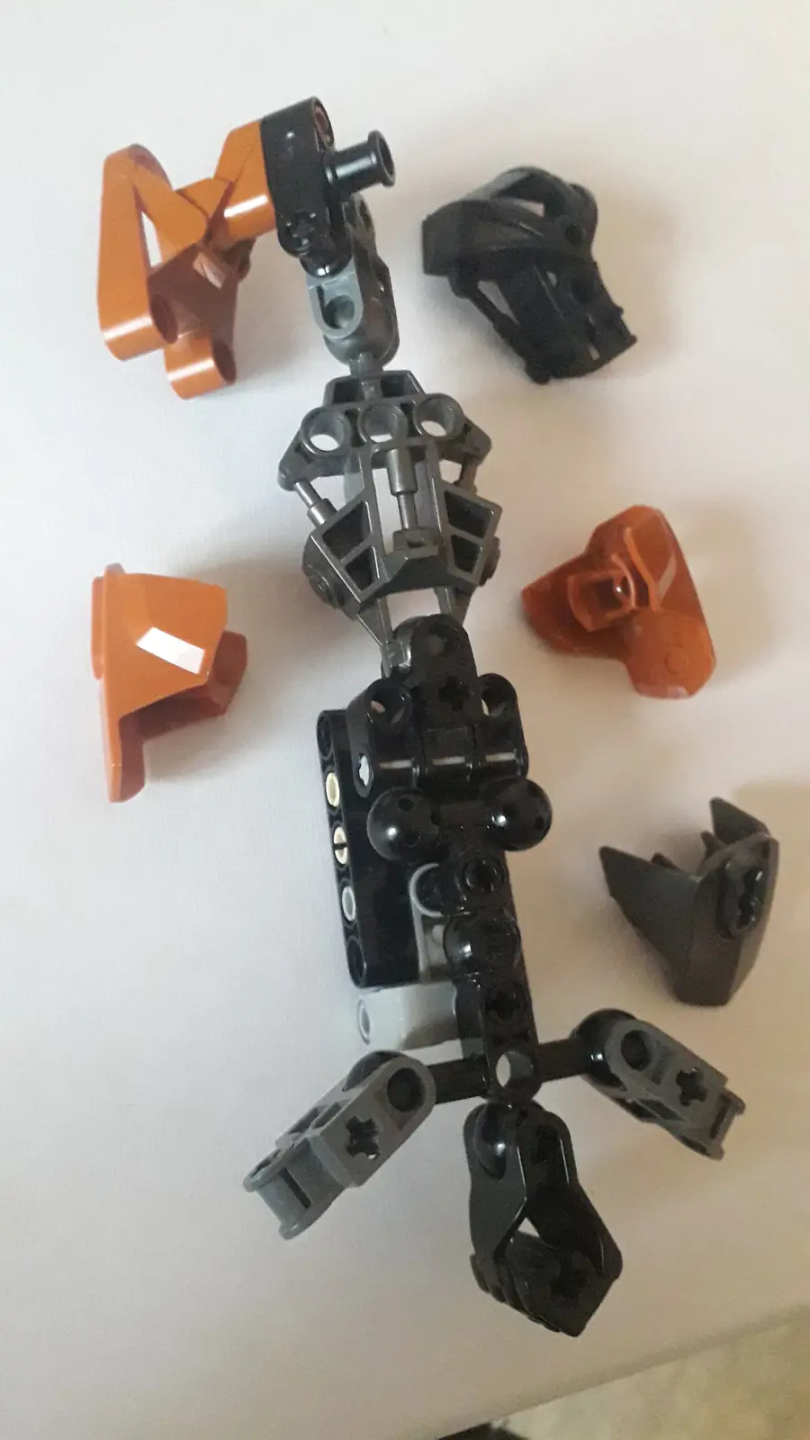 A partially assembled Bionicle-style toy figure with black and orange plastic pieces laid out on a white surface, showing a…