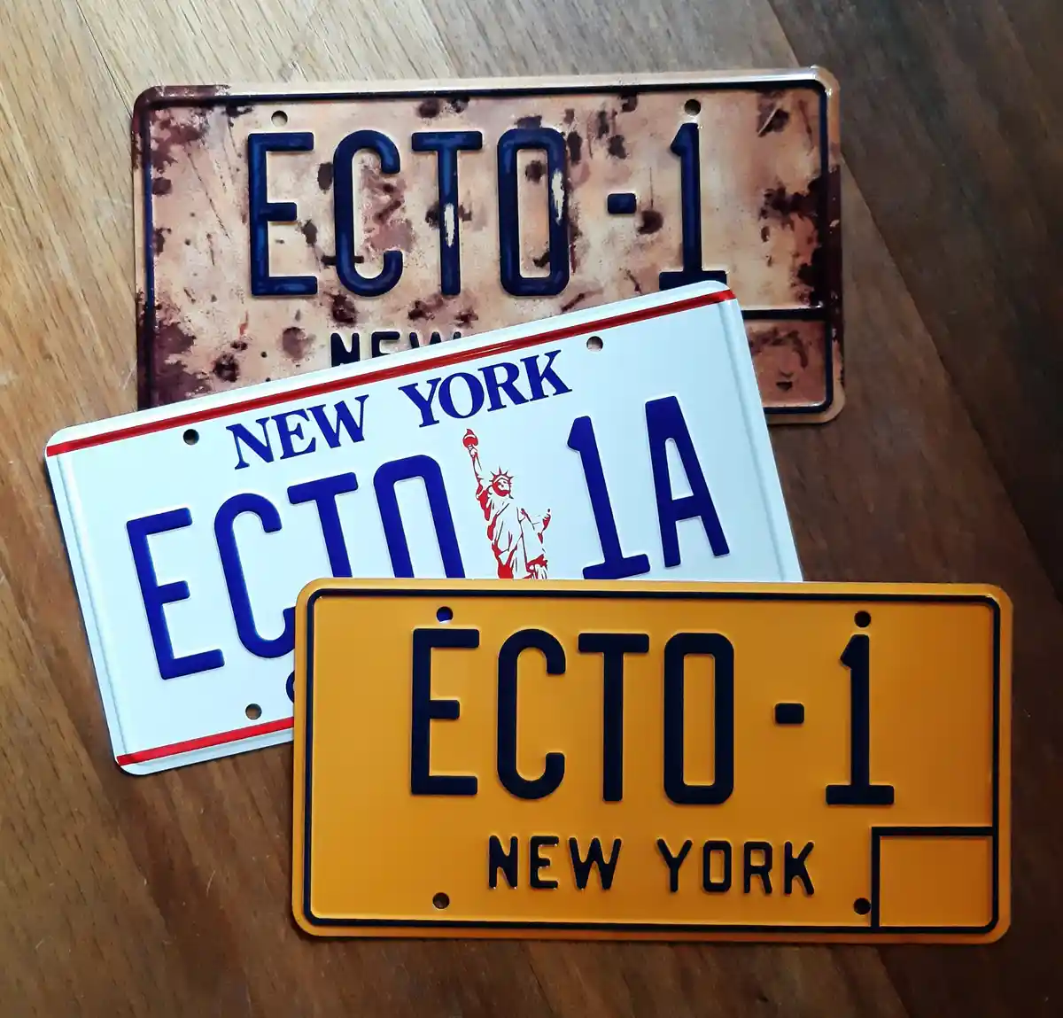 Three license plates with 'ECTO-1' are displayed on a wooden surface: a rusty brown one, a white New York plate with 'ECTO…