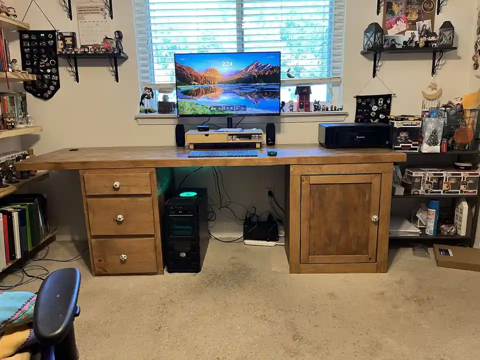 A wide wooden desk with drawers and a cabinet, holding a monitor, keyboard, and printer, surrounded by shelves filled with…