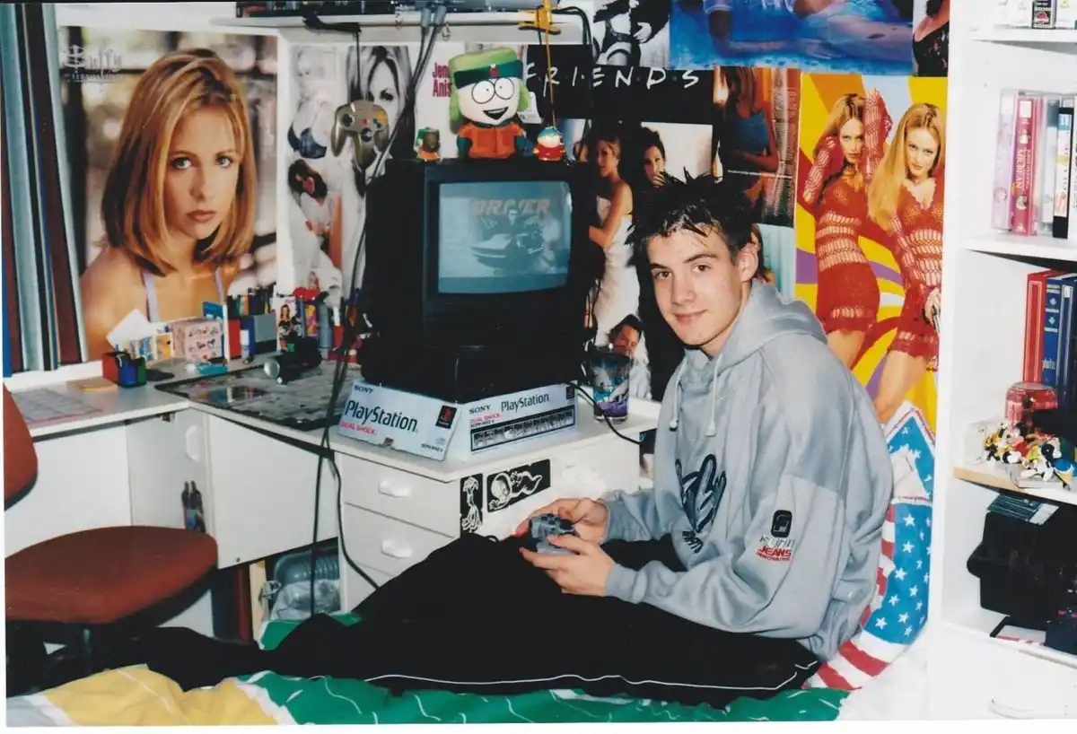 A young man sits on his bed in a cluttered bedroom, holding a PlayStation controller, with a small TV displaying a racing…