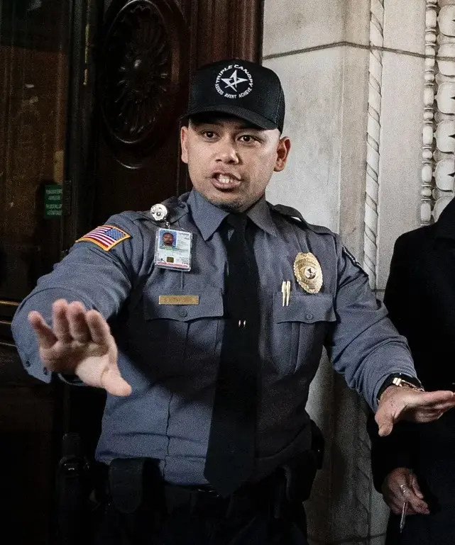 A man in a dark blue uniform with a badge, name tag, and American flag patch on his sleeve stands with both hands open…