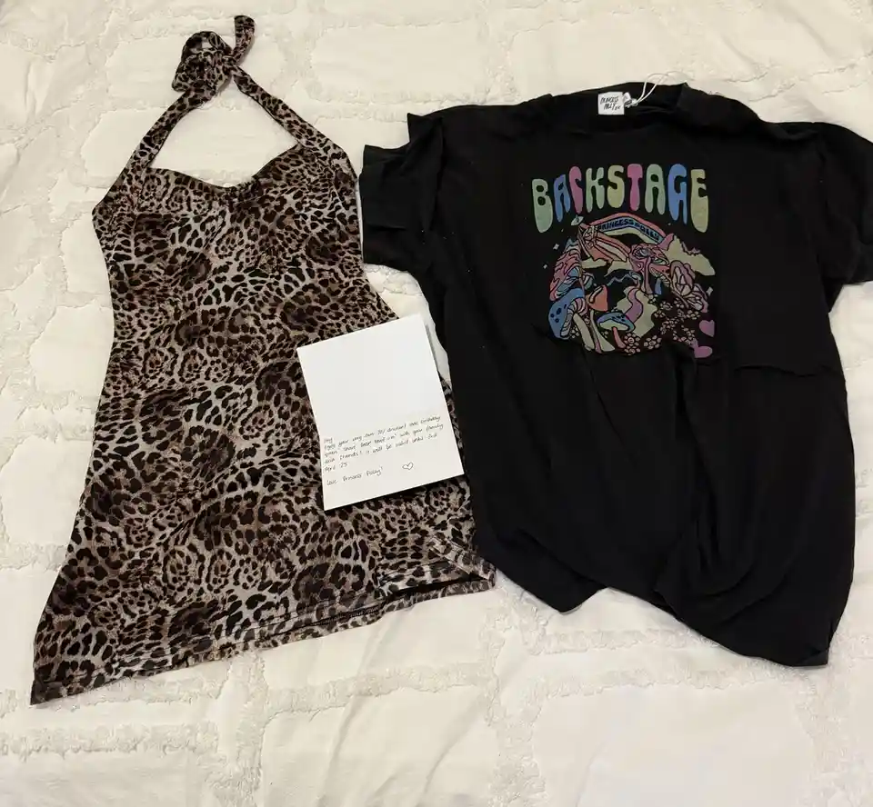 A leopard-print halter top and a black 'BACKSTAGE' t-shirt by Princess Polly lie on a white textured bedspread. A…