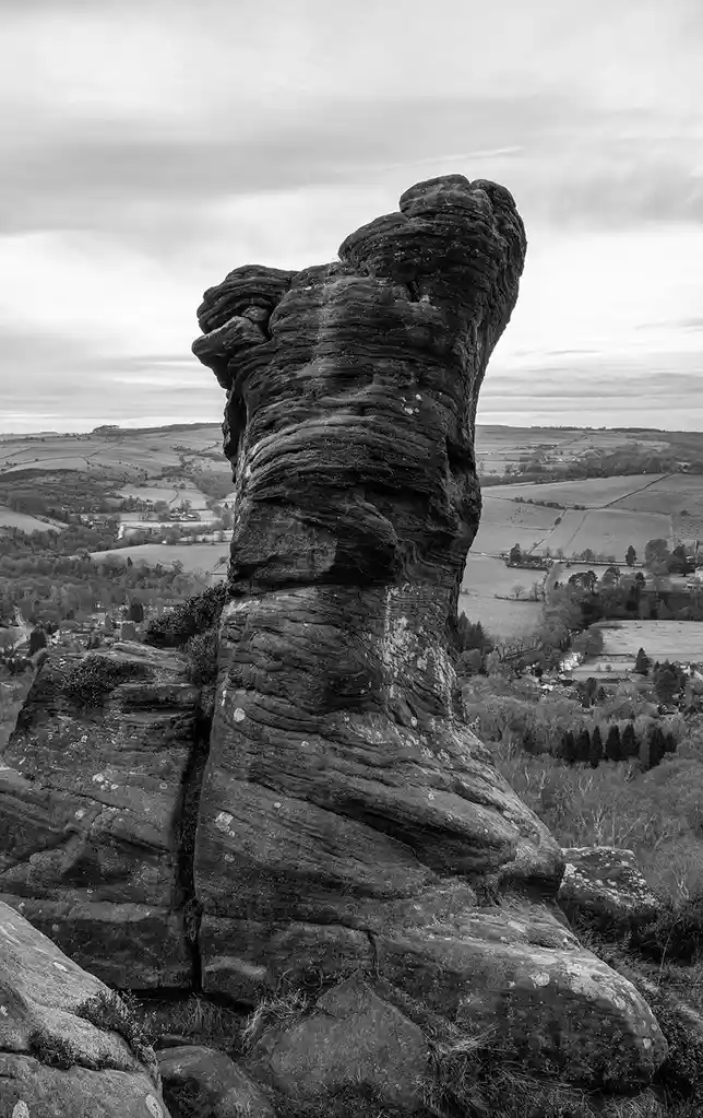 A black-and-white photograph of a tall, uniquely shaped rock formation resembling a human torso or figure, standing…