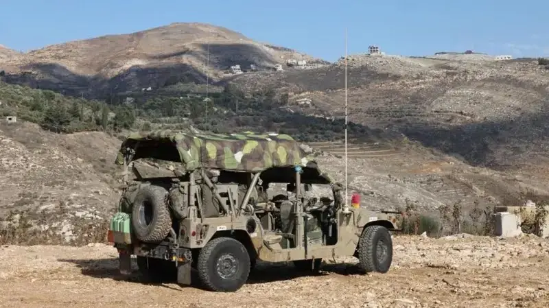 A military Humvee with a camouflage cover is parked on a dirt road in a hilly, arid landscape under a clear blue sky. The…