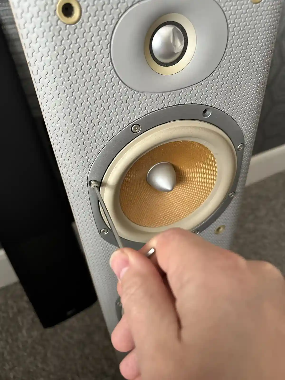 A close-up photo shows a person's hand using a small screwdriver to adjust a screw on a silver and beige speaker. The…