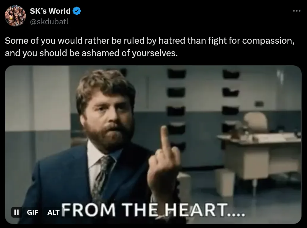A man in a suit with a beard is shown pointing his middle finger directly at the viewer while standing in an office-like…