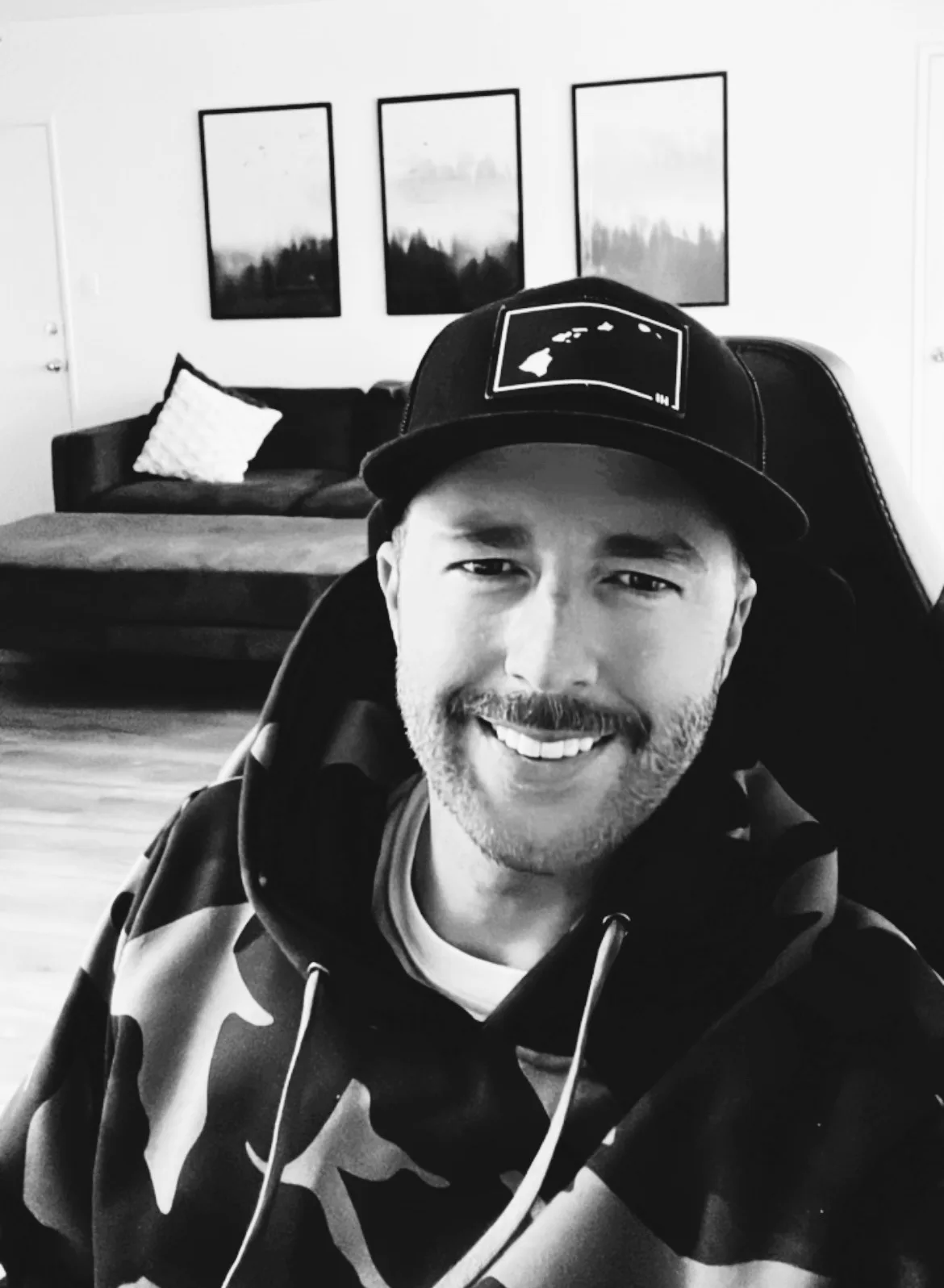 A black-and-white photo of a smiling man with a beard wearing a camouflage hoodie and a black baseball cap with a white…