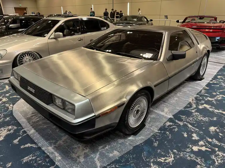 A silver DeLorean DMC-12, famous from 'Back to the Future,' is displayed at a car show on a patterned blue carpet…